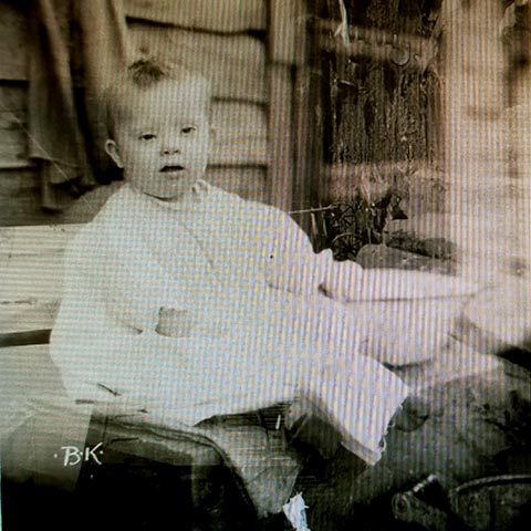 A baby with Down Syndrome sits on a bench outside a house, next to an old-fashioned wash basin.