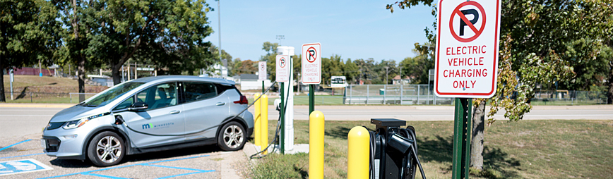 A State of Minnesota fleet vehicle is plugged into a charger in the background while a sign in the foreground says 'electric vehicle charging only'