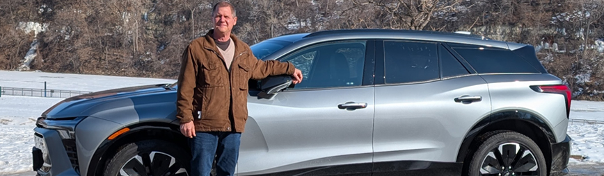 an older man stands in front of his electric vehicle parked in front of a snowy landscape