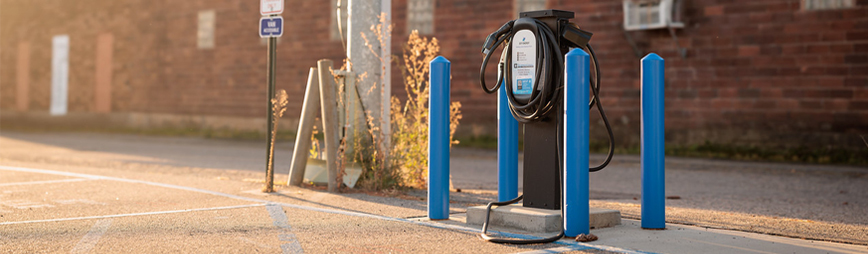 a dual-port level 2 EV charger surrounded by blue bollards in a parking lot as the sun sets.