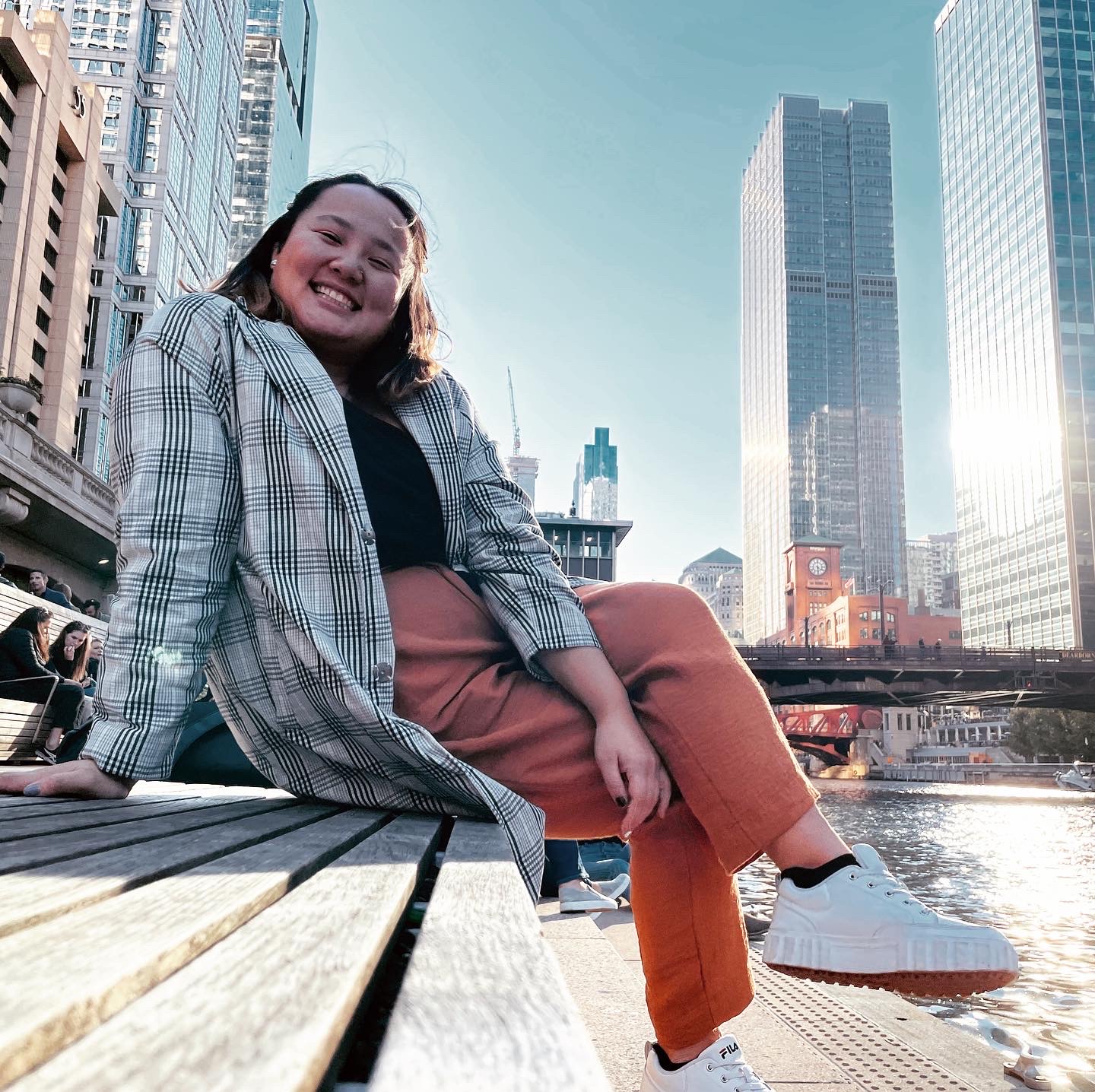 A snapshot of Makayla grinning as she poses, sitting on a bench with a city skyline behind her. The camera view is pointing up at her.