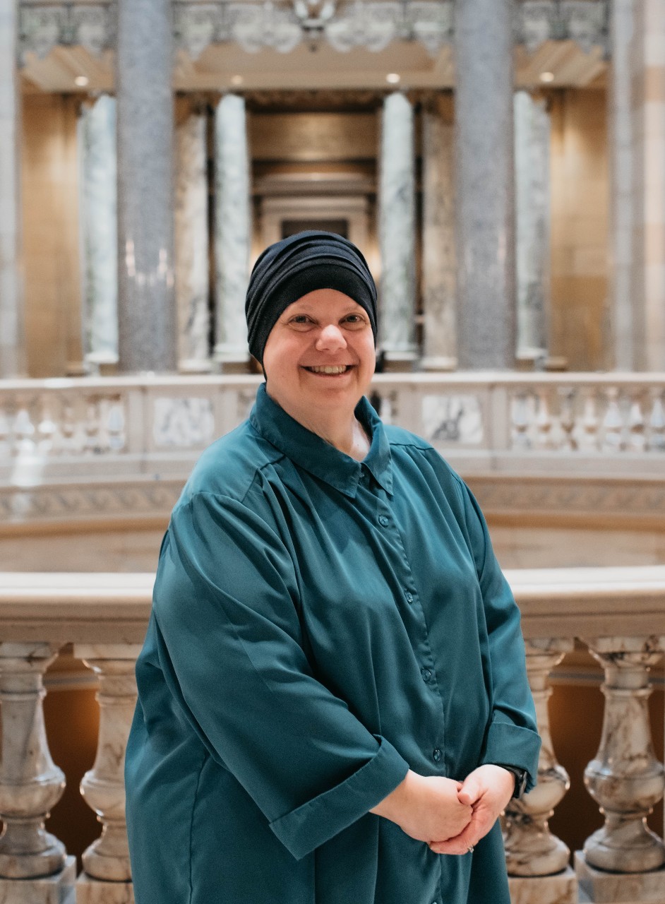Becky is wearing a teal business shirt. She is smiling. Behind her is the interior Rotunda inside the Capitol.