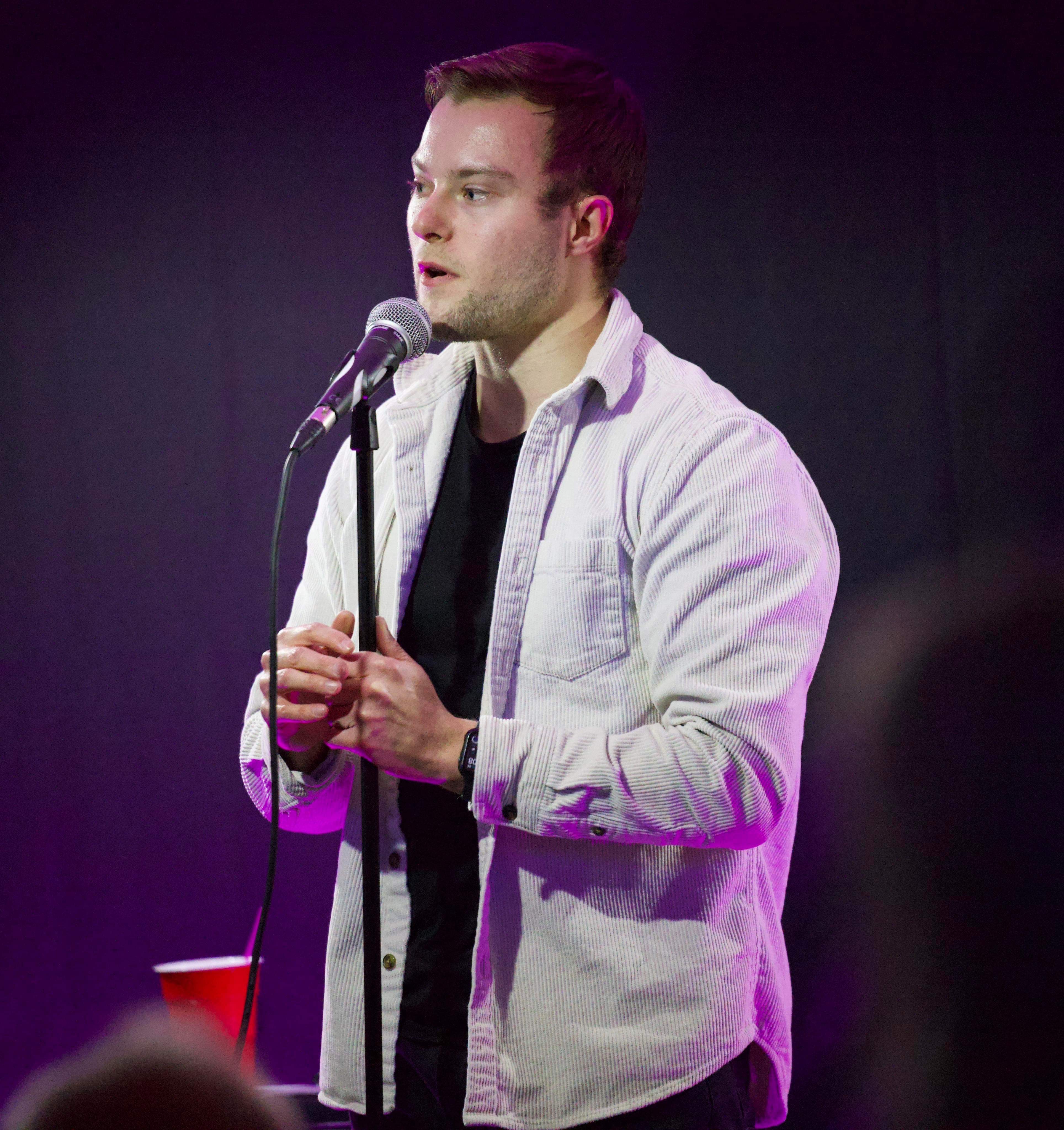 Snapshot of Sam standing onstage wearing an open white shirt and holding his hands together. In front of him is a standing  microphone.