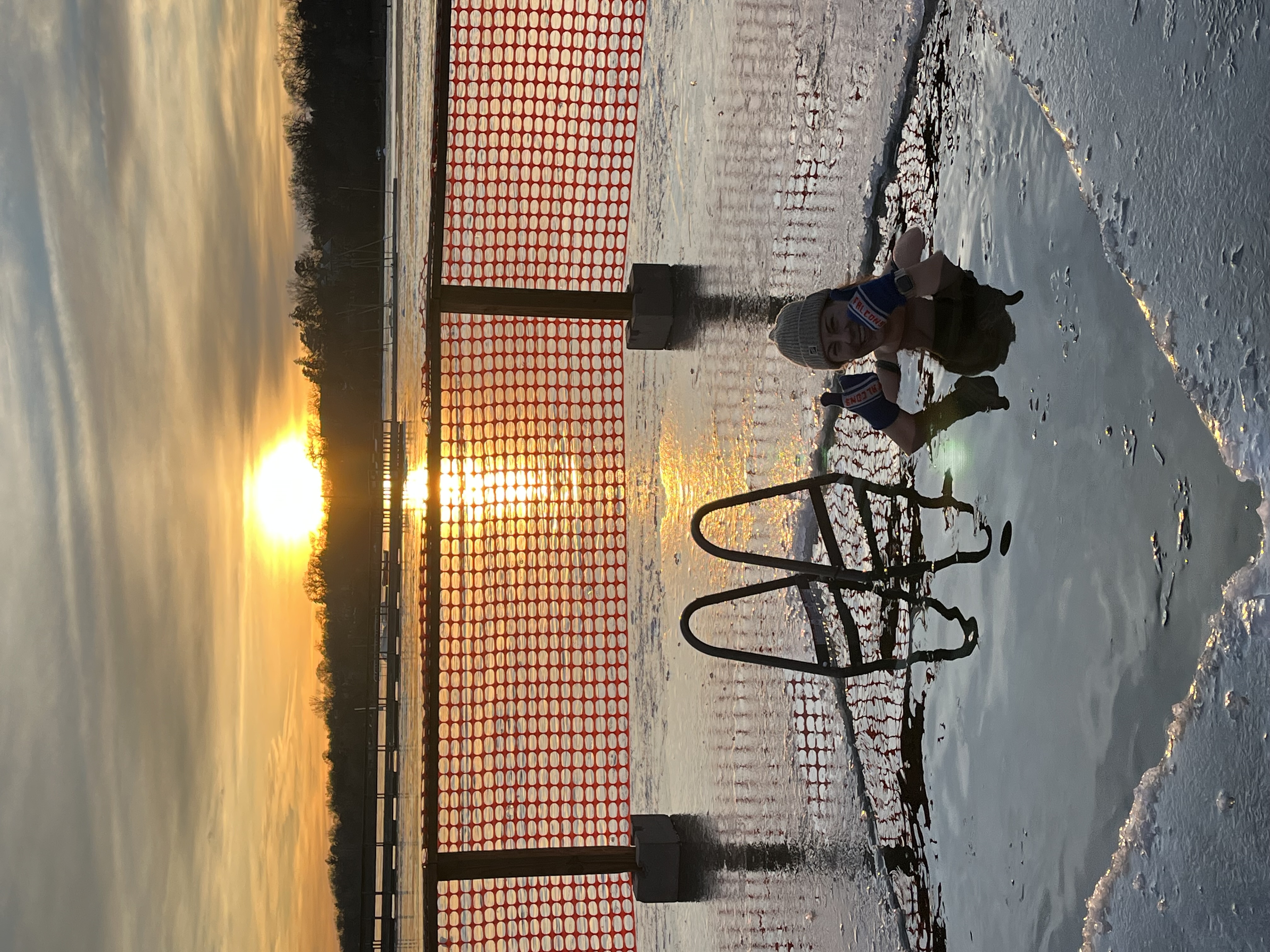 Haley in silhouette, in a chest-high water hole cut out from the ice standing next to a ladder, with a lakeside sunset landscape behind her.