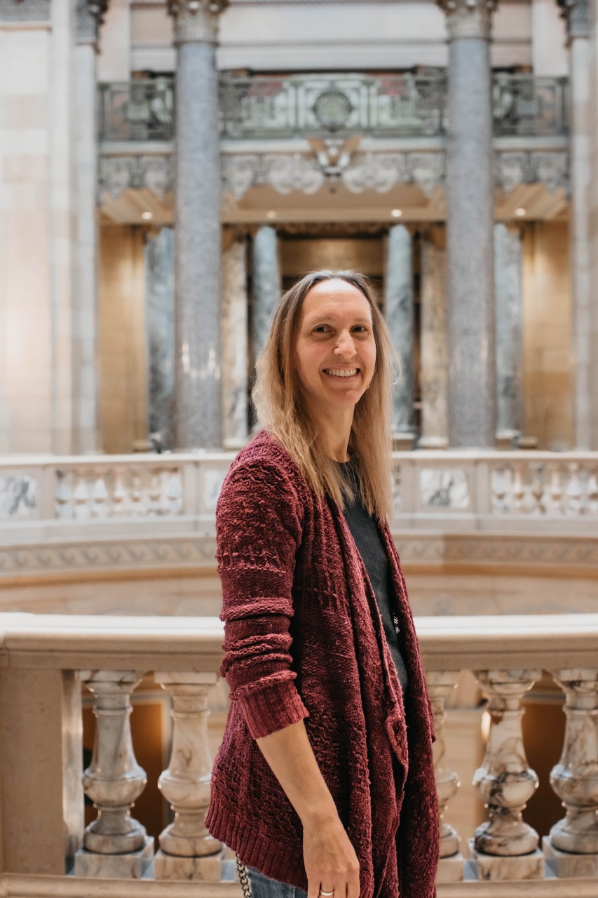 Krista has long brown hair. She is smiling and standing inside the Capitol with the Rotunda behind her.