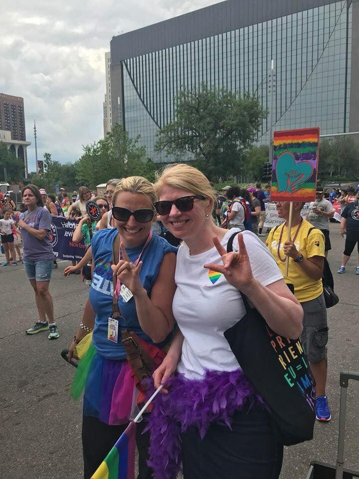 Krissy and Jessalyn walking among folks at the Pride festival. They both are wearing sunglasses, colorful Pride gear, and Krissy carries a flag in one hand. They both flash the ‘I love you’ sign to the camera.