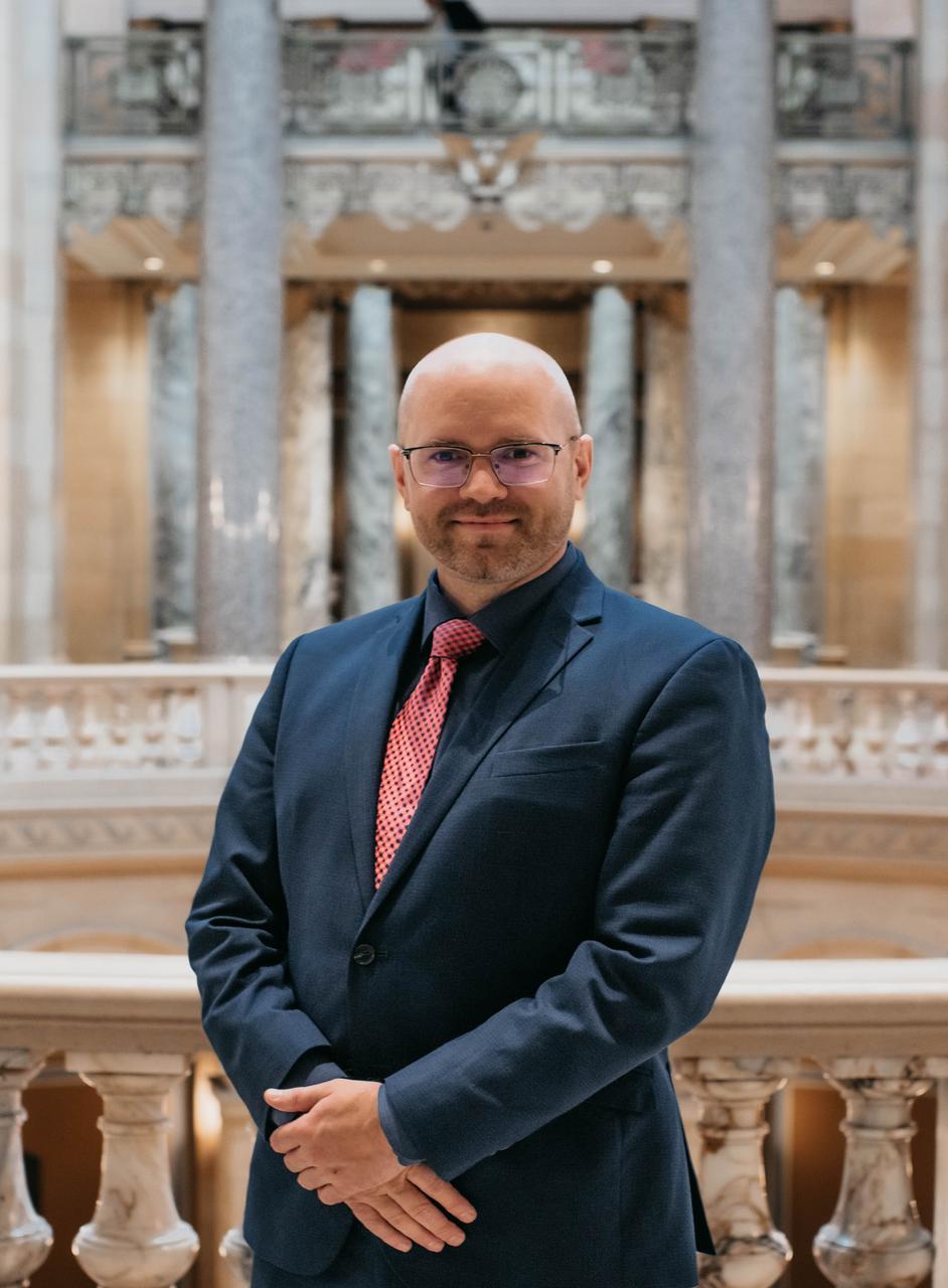Keith is wearing a business suit. He is standing inside the Capitol with the Rotunda in the background.