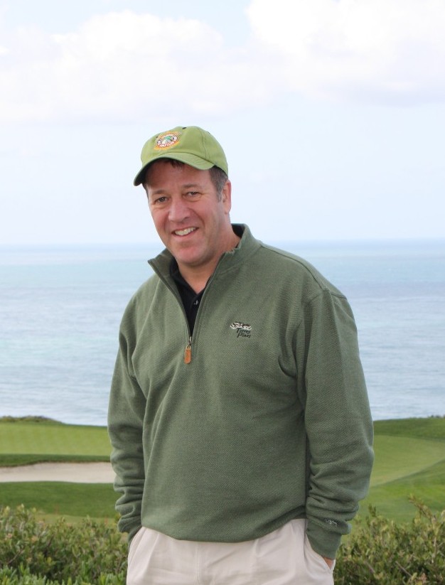 Casual snapshot of Mark, wearing outdoorsy clothes and hat, hands in pockets and grinning as he stands outside on a golf course.