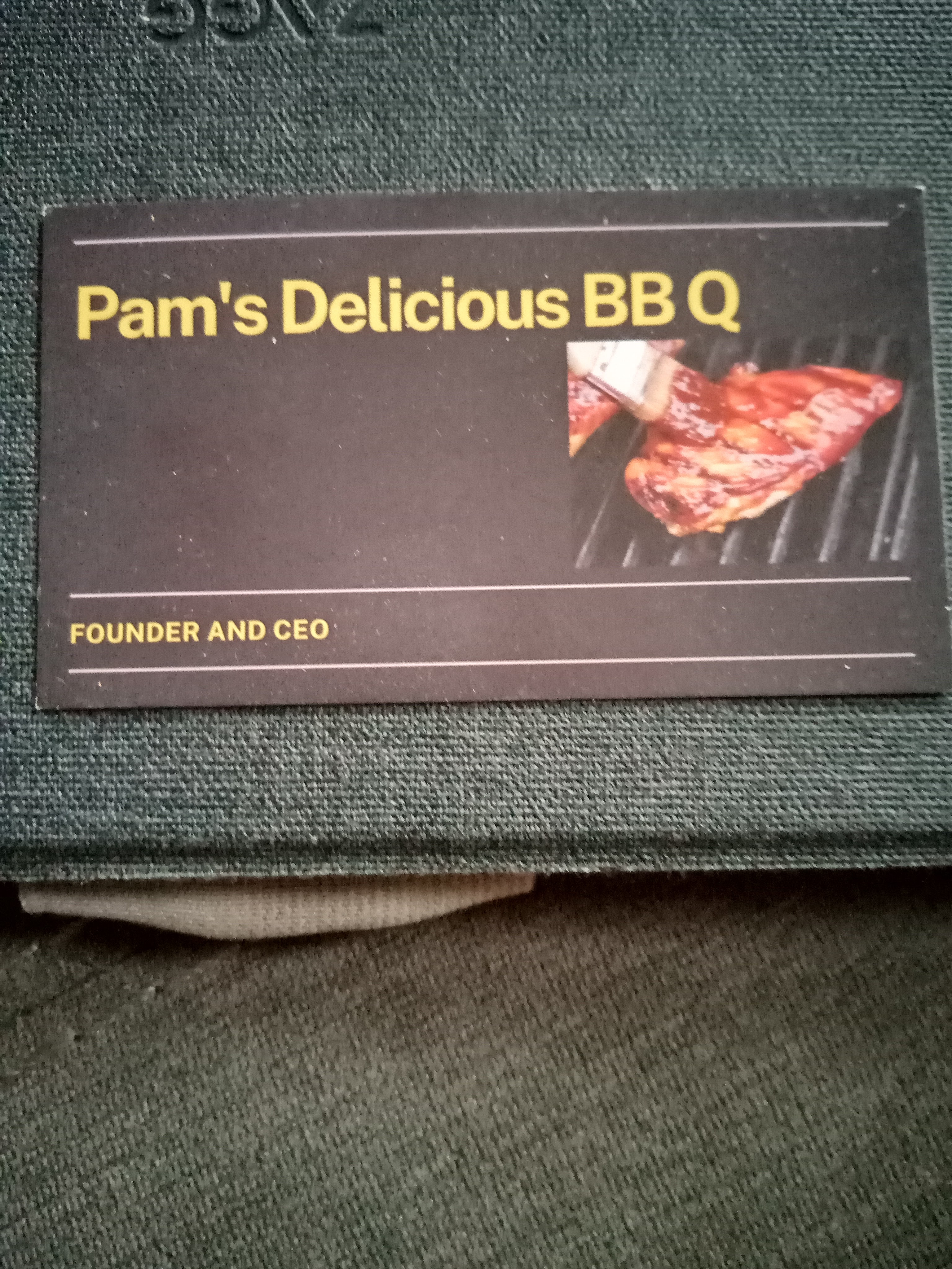 Close-up of a dark-colored business card with gold text and lines. Text across the top: “Pam’s Delicious BBQ”, with “Founder and CEO” across the bottom. A photo of a BBQ-sauce slathered meat on a grill is on the right side.