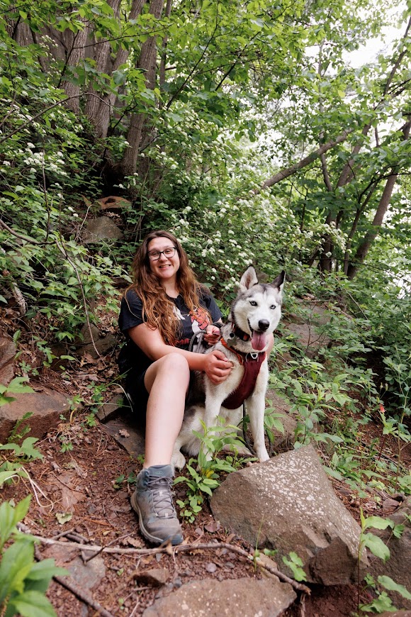 Sophia is wearing a black Green Day t-shirt smiling at the camera with her dog in front of her. Her dog is a Siberian Husky with gray and white fur. They are sitting on a pile of rocks at a hiking trail. Surrounded by trees.