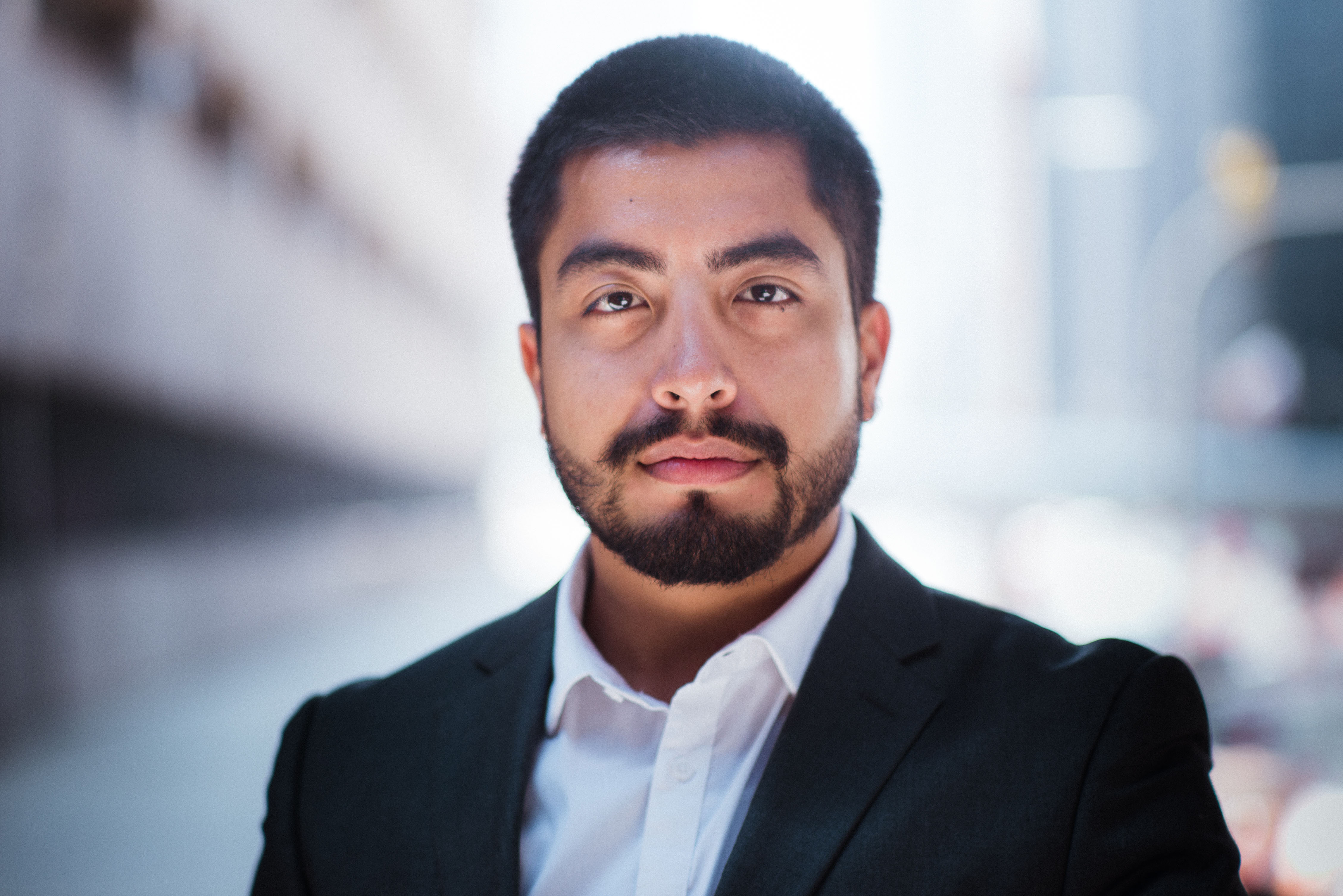 A man faces the camera with a serious facial expression. He has a beard, mustache, and is wearing business attire.