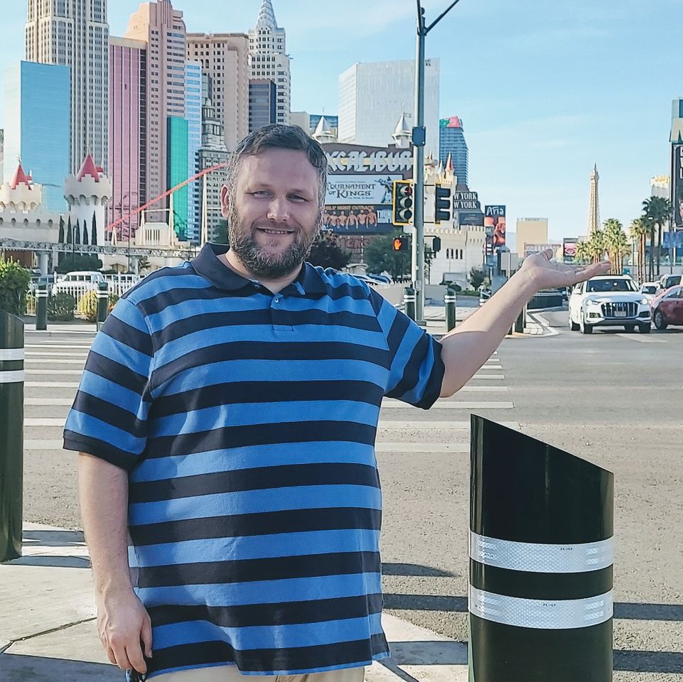 Jeremy stands on a street in Las Vegas, standing next to a black and white striped bollard. Jeremy is wearing a blue striped shirt and posing, showing off the buildings behind him.