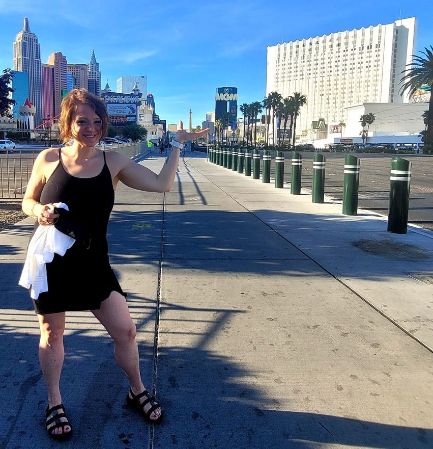 Jessi, wearing a black sundress and holding a scarf in one hand, grins as she poses on a wide boulevard in Las Vegas, one arm stretched out to the side, hand facing up and flat. A small tower in the distance looks like it is balancing in the palm of her hand. Other buildings fill the background with a row of bollards separating the boulevard from the road running alongside it.