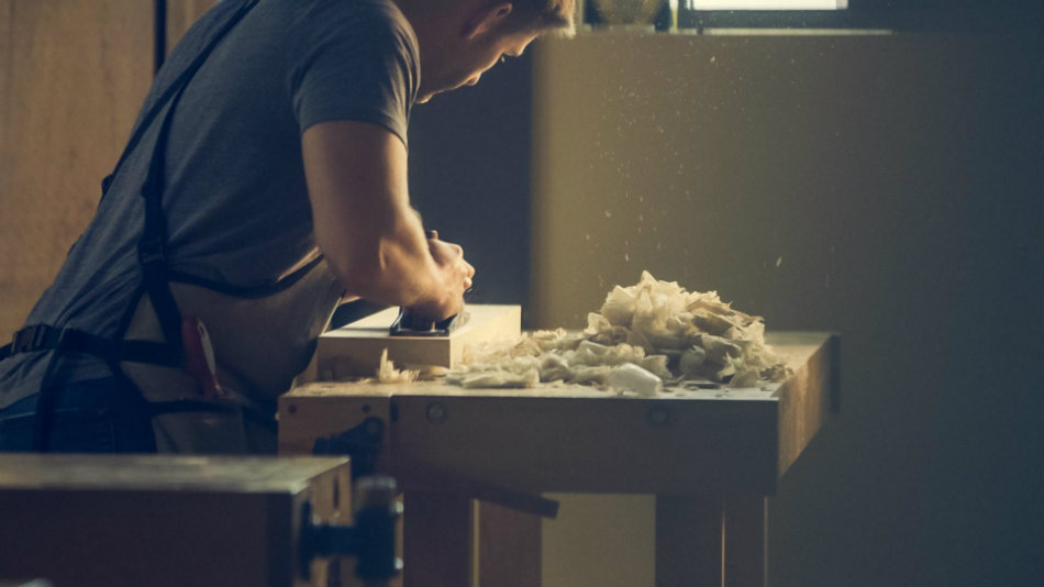 A man is at his workbench He is sanding a wooden board. 
