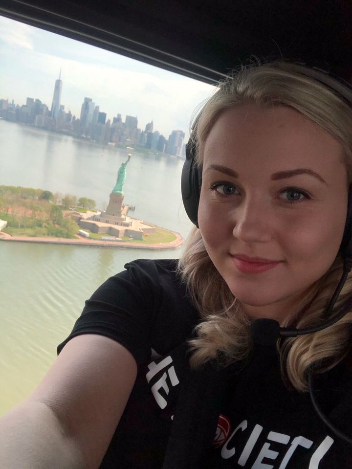Kaylah, a white woman with long wavy blonde hair and wearing a black shirt with white text across the chest, and headphones on her ears, smiles as she sits inside a helicopter. Behind her is a bird’s eye view of the Statue of Liberty in the harbor of New York City, with the city skyline in the distance.