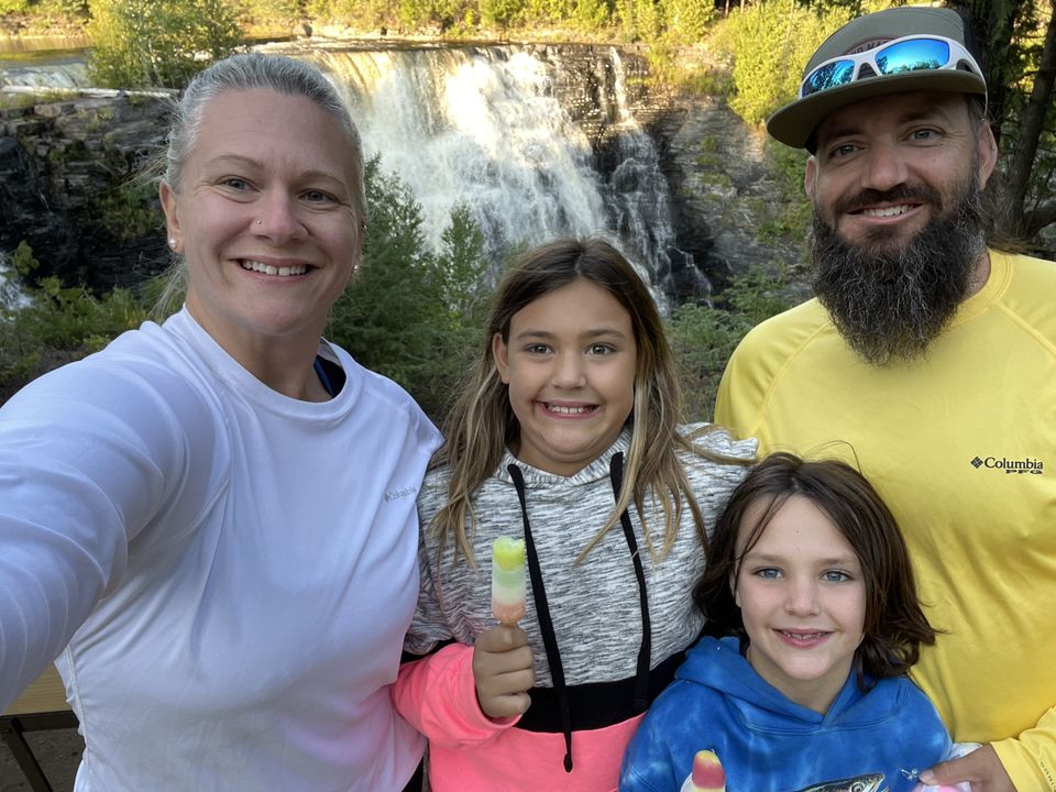 Marissa poses with her family in a group snapshot standing outside with other people visible in the background. She stands next to two children and a man.
