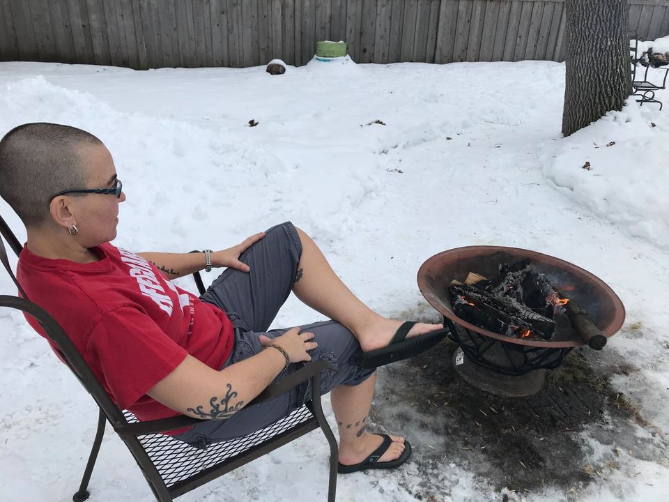 Nic relaxes in a patio chair in the backyard, watching a smoldering fire in the firepit in front of her. There is still snow on the ground.