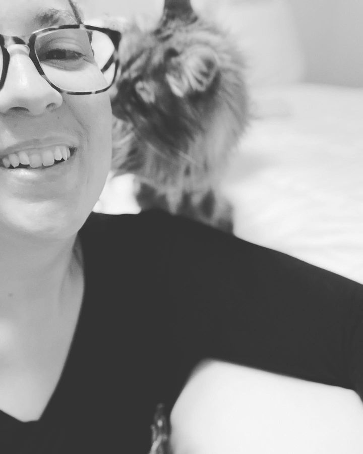 Sammie smiles forward to the camera. She wears a black long sleeve and her right arm is propped up on the futon. Her cat, Sue, is on top of her right shoulder and gives Sammie a kiss on her cheek.