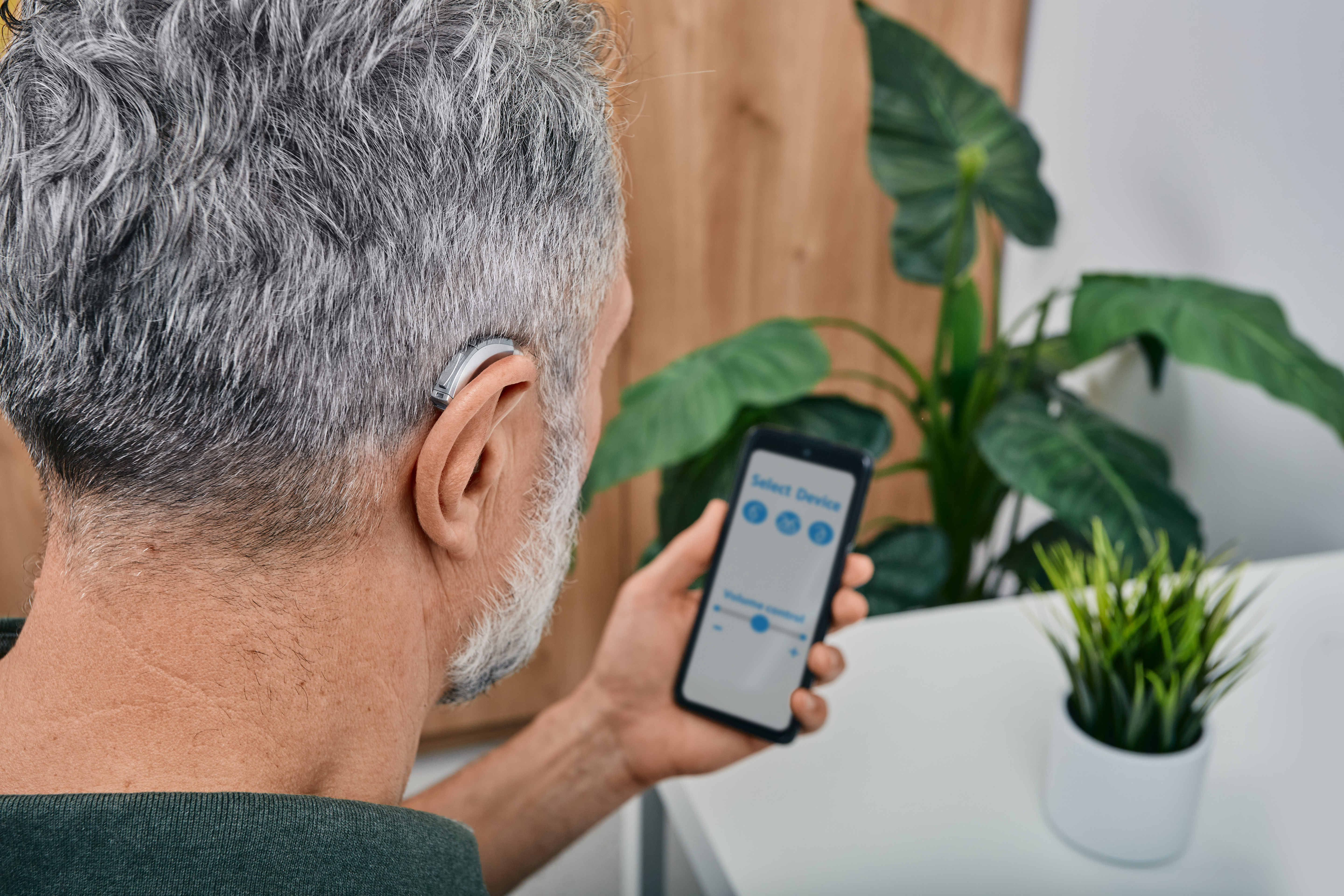 Man with silver hair adjusting his hearing aid with his smartphone.