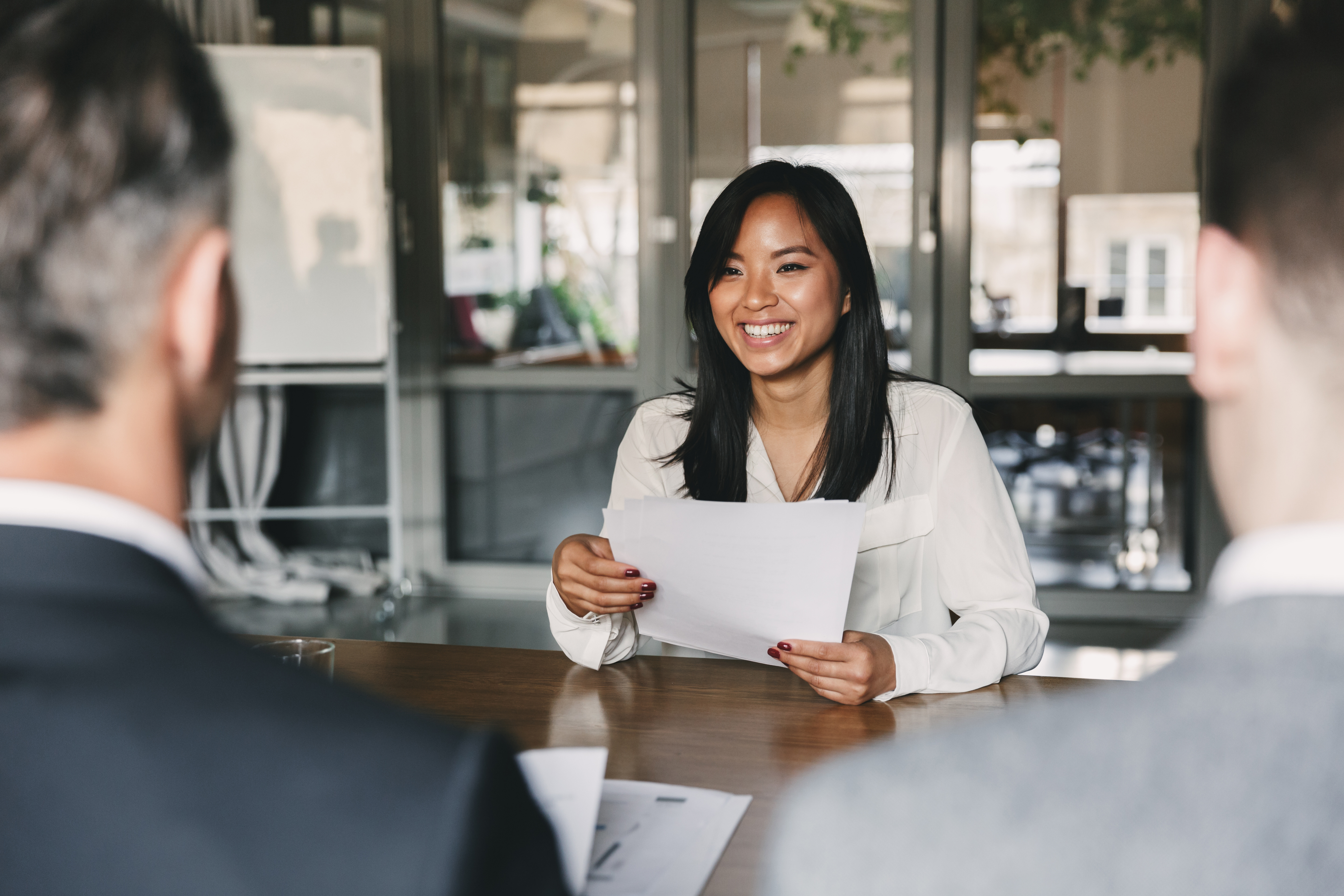 A woman with straight black hair and brown skin holding a sheet of paper and smiling at her interviewers. 