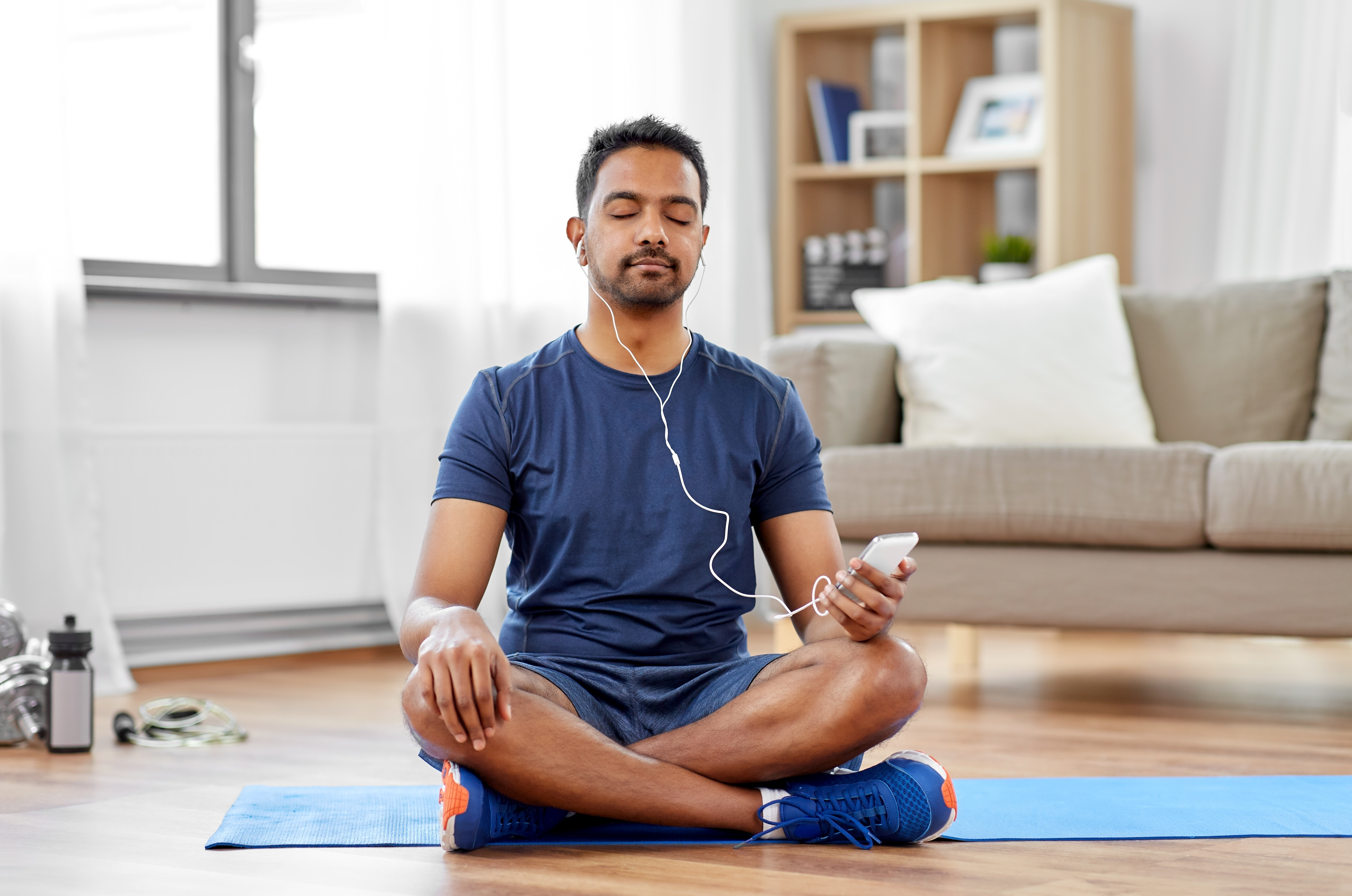 A man sitting, legs crossed, on an exercise mat with his eyes closed. He is wearing earphones and holding his smartphone in his hand.