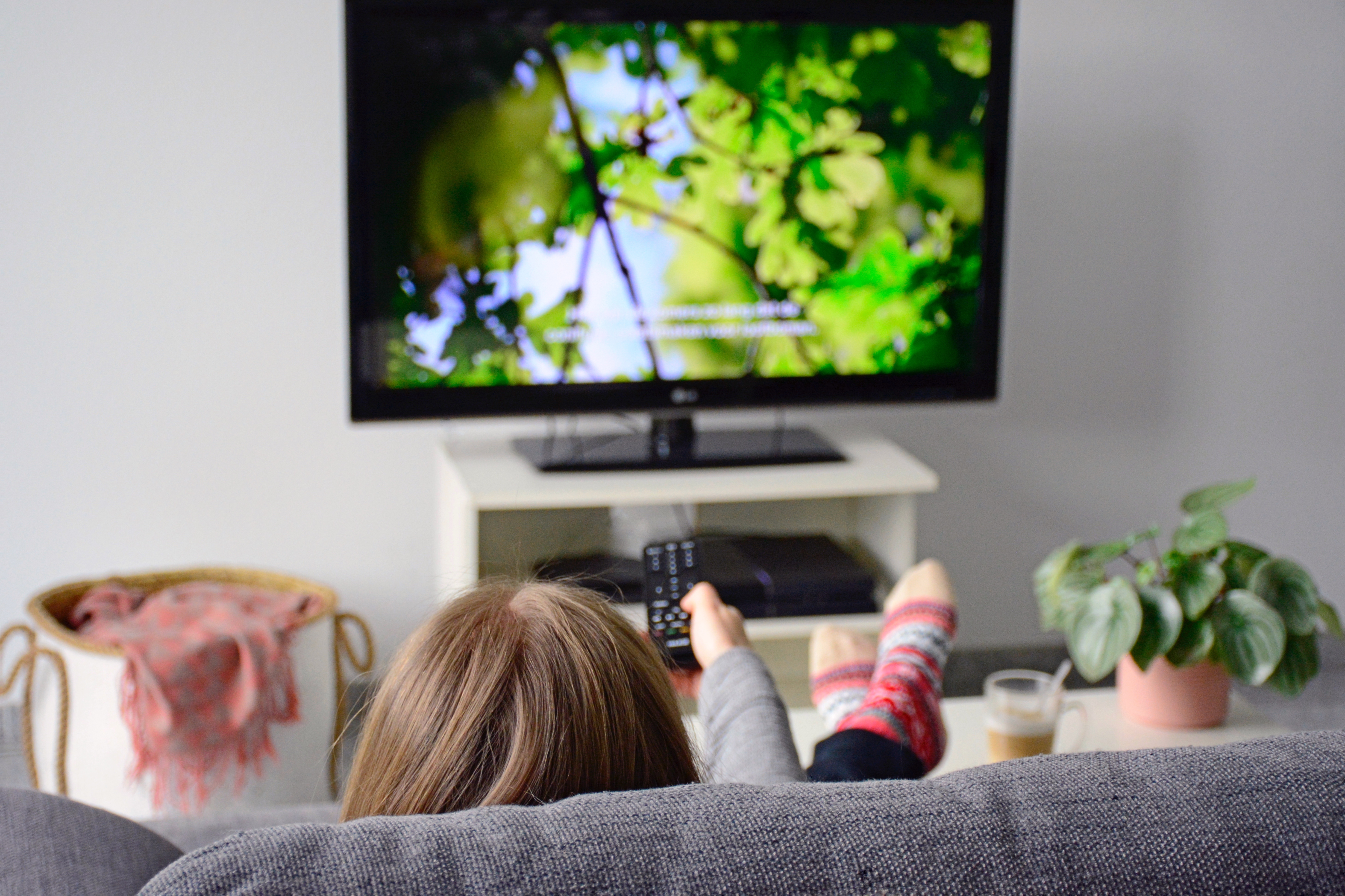 A person is sitting on a couch with a remote pointed at a television screen showing green trees and captions. 