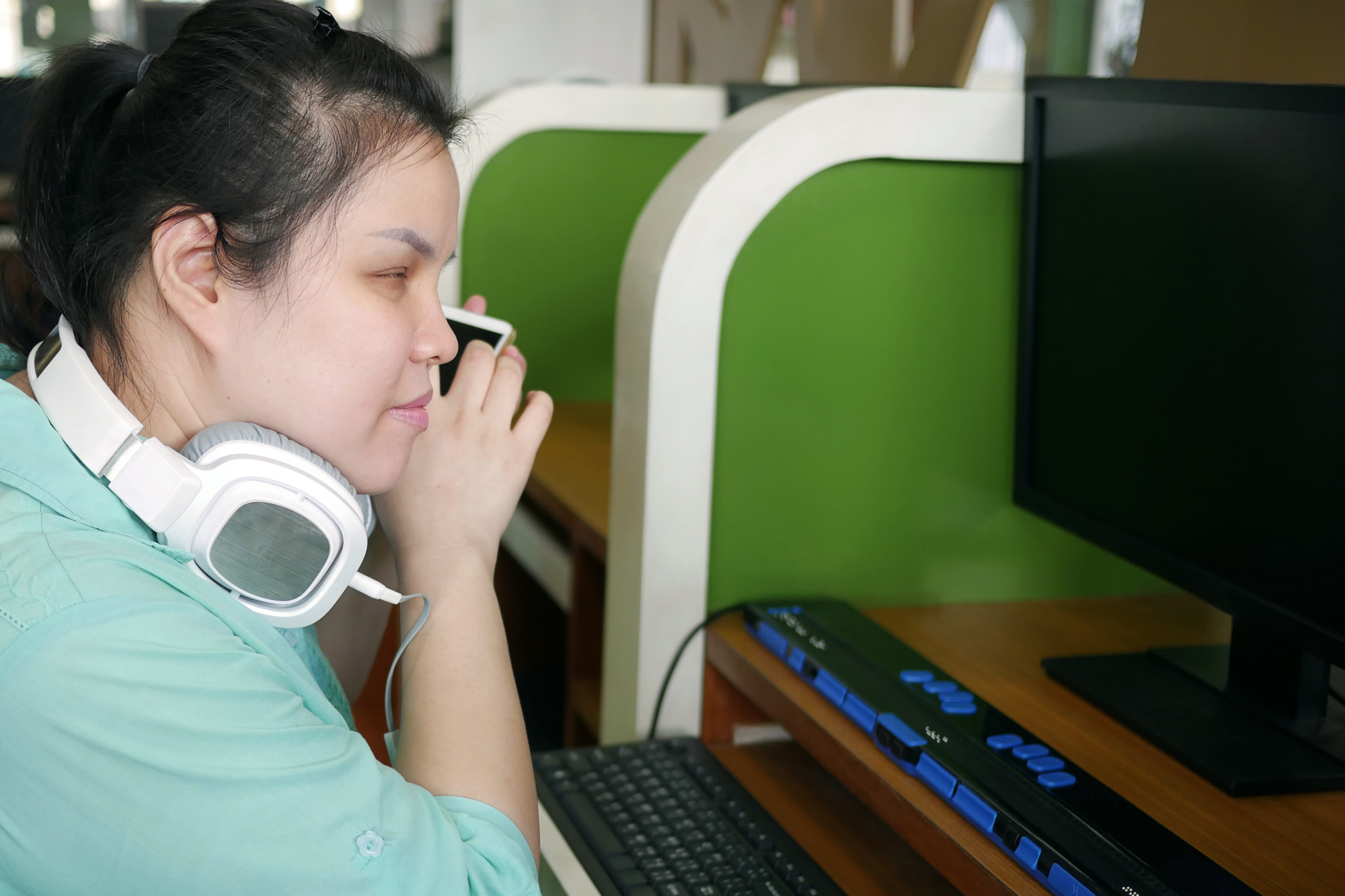 Woman with vision loss at computer with braille display. She has earmuff style headphones around her neck and is holding her smartphone up close to her ear.