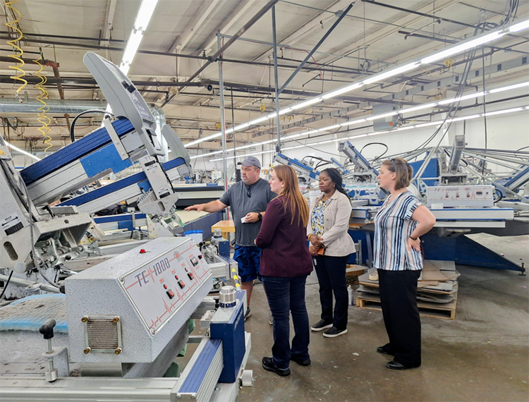 Five people stand beside a large machine as a man describes how it works.
