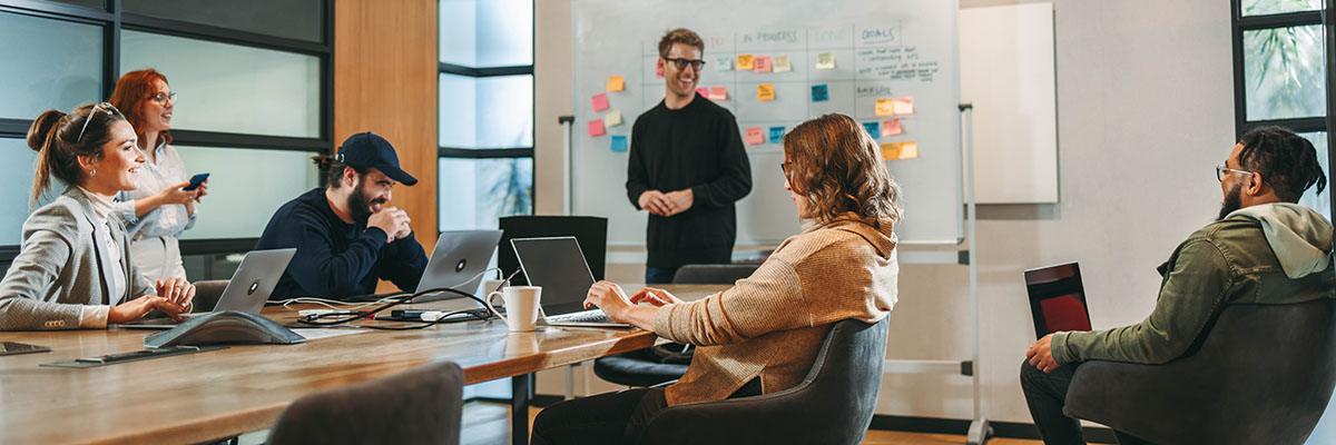 A group of six people gathered around a desk with laptops in a conference room brainstorming ideas with Post-It Notes on a whiteboard A group of six people gathered around a desk with laptops in a conference room brainstorming ideas with Post-It Notes on a whiteboard