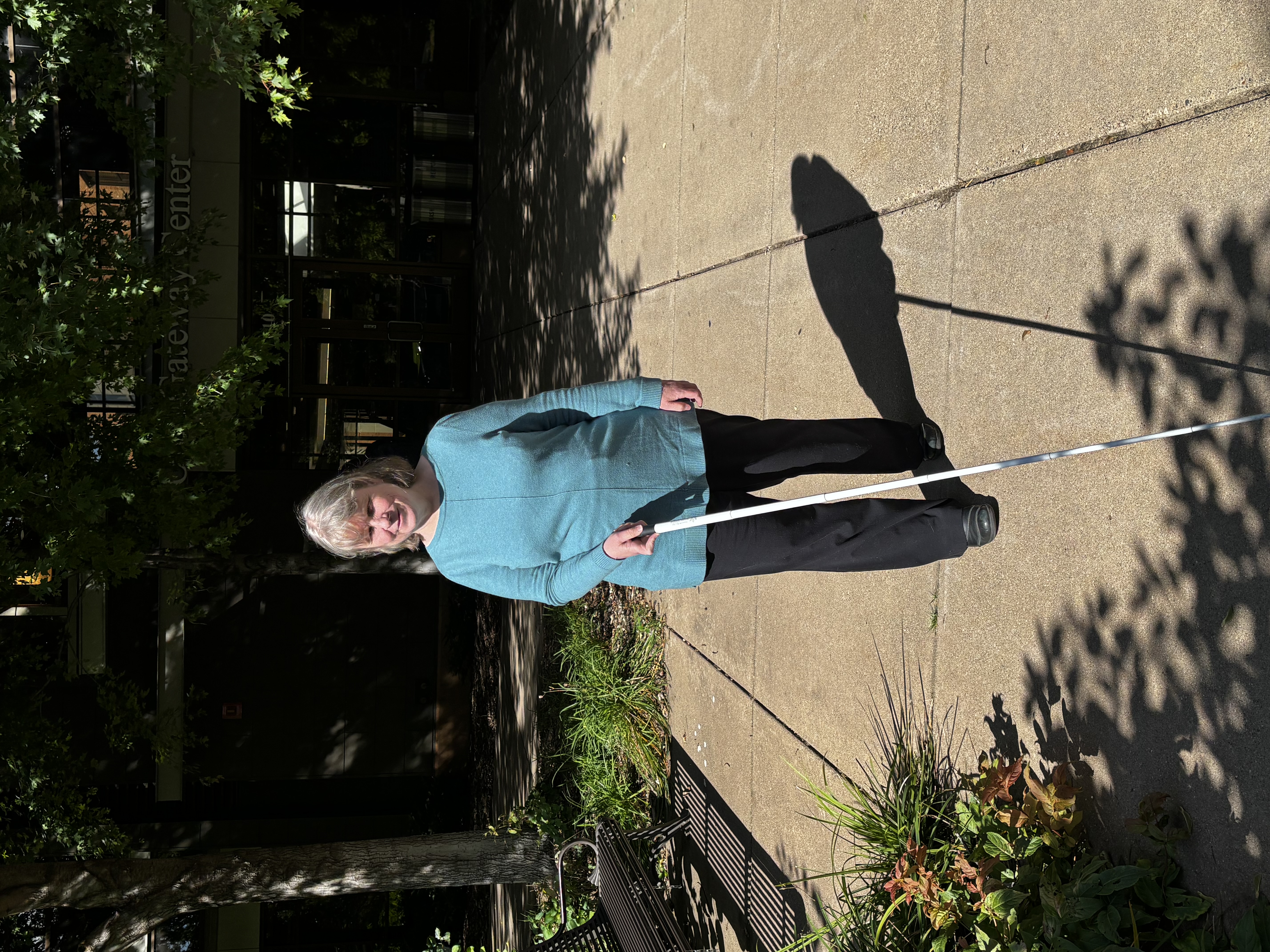 Kathy walking with her white cane in front of her. She's wearing a teal sweater and black pants. She's in front of the Oren Gateway Center on the Augsburg University campus.