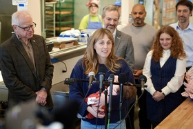 The owner of Marty's Deli stands in front of a microphone with her baby in her arms. Governor Walz and Commissioner Varilek stand behind her.