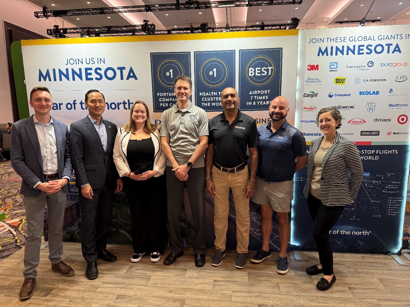 A group of 7 people standing in front of Minnesota's booth at RE+. The booth backdrop features Minnesota's accolades as number 1 in Fortune 500 companies per capita, number 1 health tech cluster in the world, best airport 7 times in 8 years, and lists companies headquartered in Minnesota.