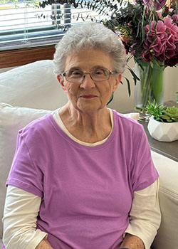 A smiling grandma sits in her living room with flowers and succulents behind her.