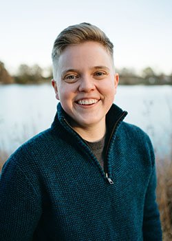 A photo of Luke Daily wearing a dark blue sweater with a zipper and a black shirt underneath. He has dark blond hair and a friendly smile.