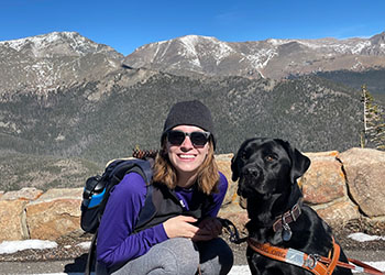 A woman smiling brightly in front of a mountain range with her guide dog.