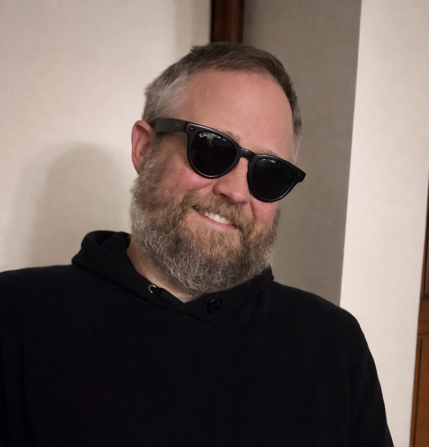 A smiling man wearing dark sunglasses and a black hoodie