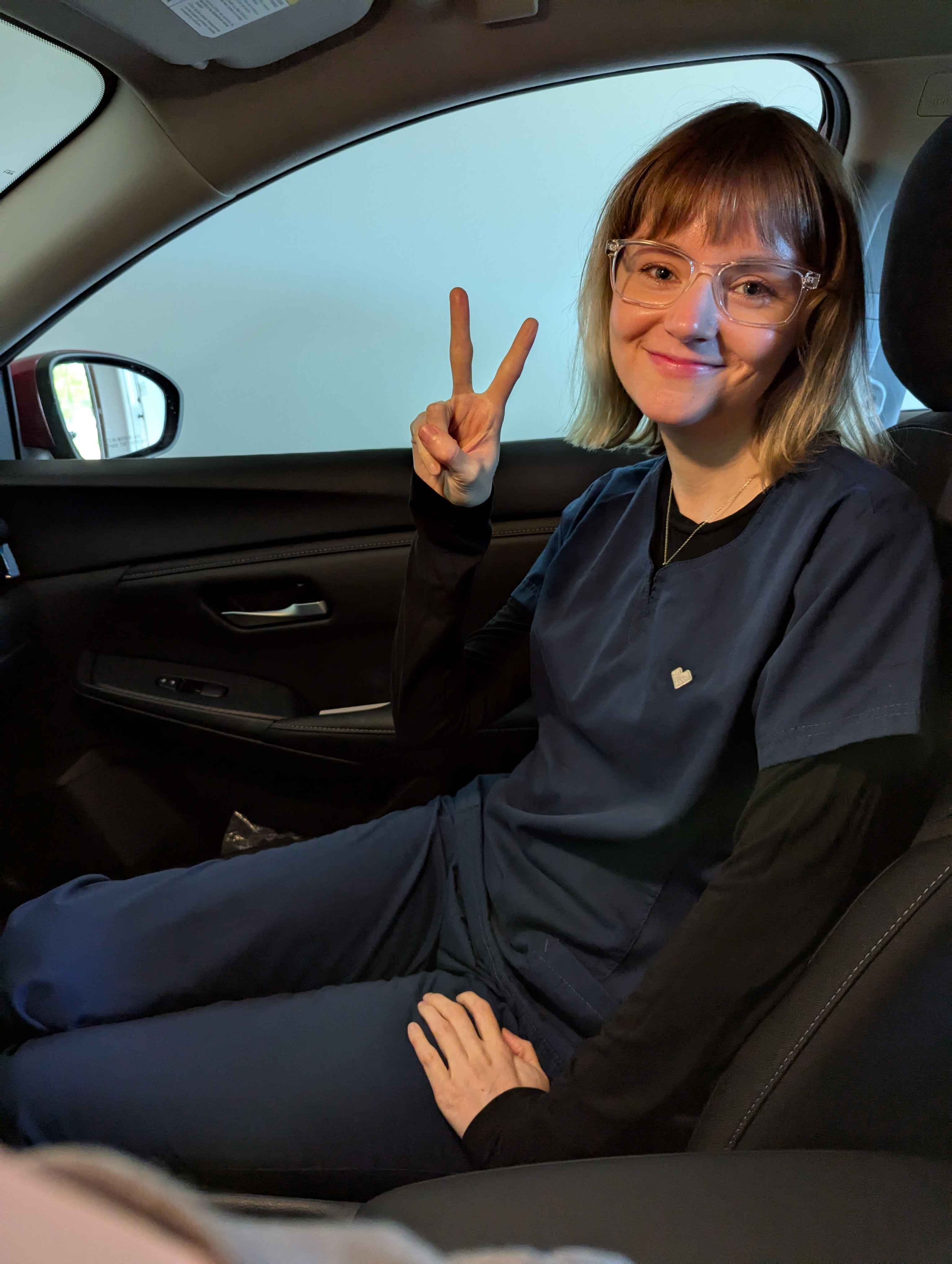 A woman in a CVS scrubs does a peace sign pose in a passenger seat
