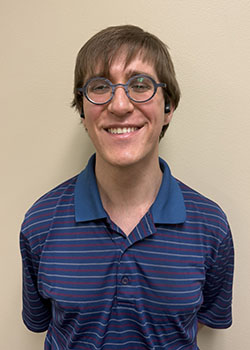 A photo of Tristan Ratliff wearing a blue polo shirt with red and light blue horizontal stripes. He has short brown hair and round glasses. He is smiling at the camera.