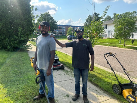 Two men holding lawn care equipment smile for a picture.