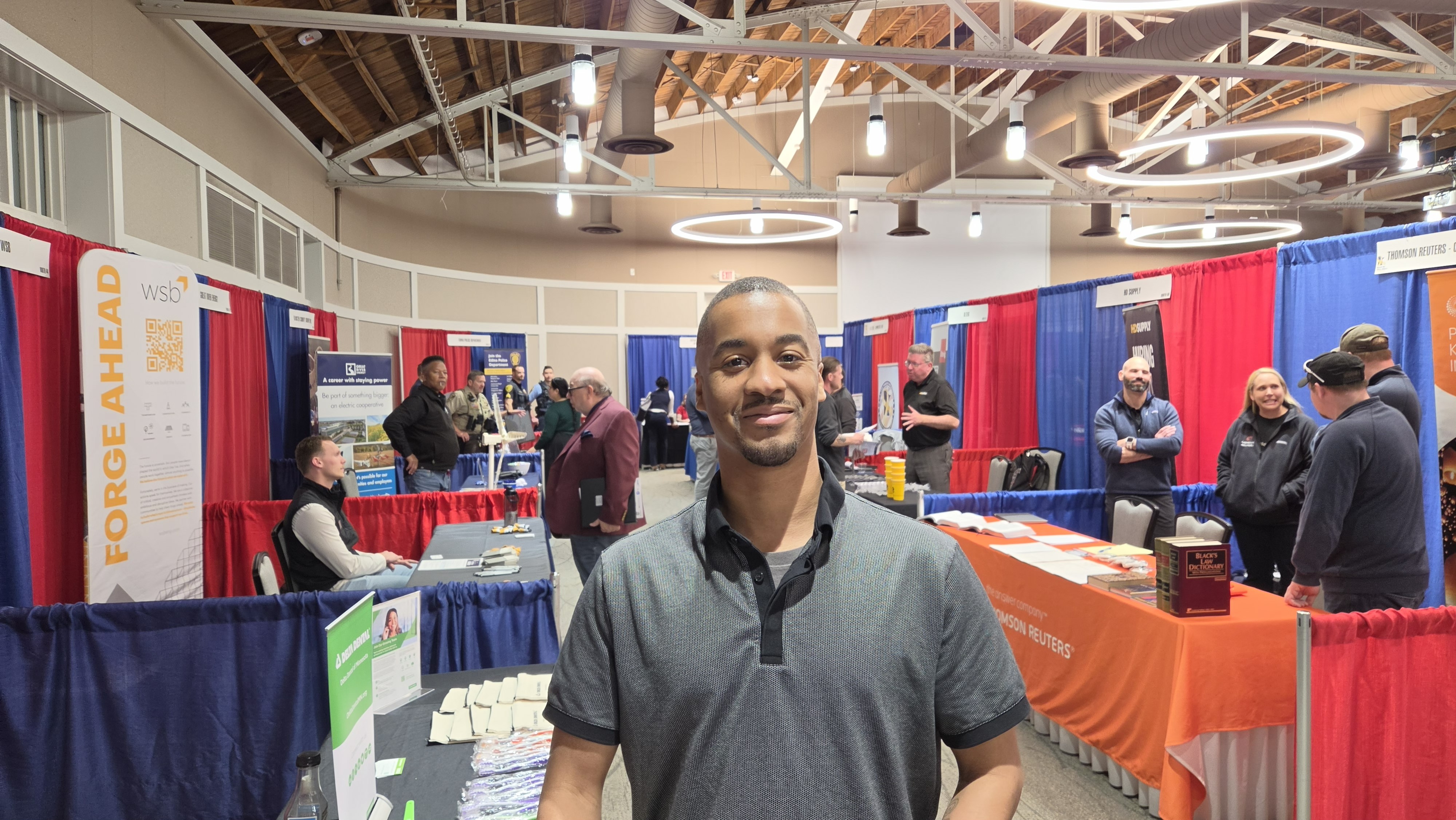 Theodore stands in the middle of the aisle at the career fair, smiling for the picture. Around him are booths set up with employers behind them, draped in red and blue. Theodore stands in the middle of the aisle at the career fair, smiling for the picture. Around him are booths set up with employers behind them, draped in red and blue.