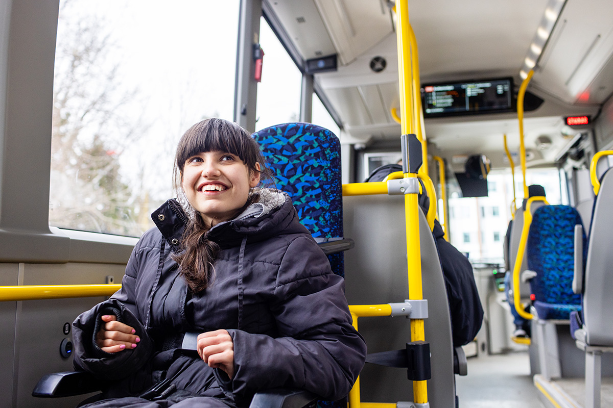 Disabled young woman in a winter coat sits on a city bus with seat belt buckled. She is smiling.