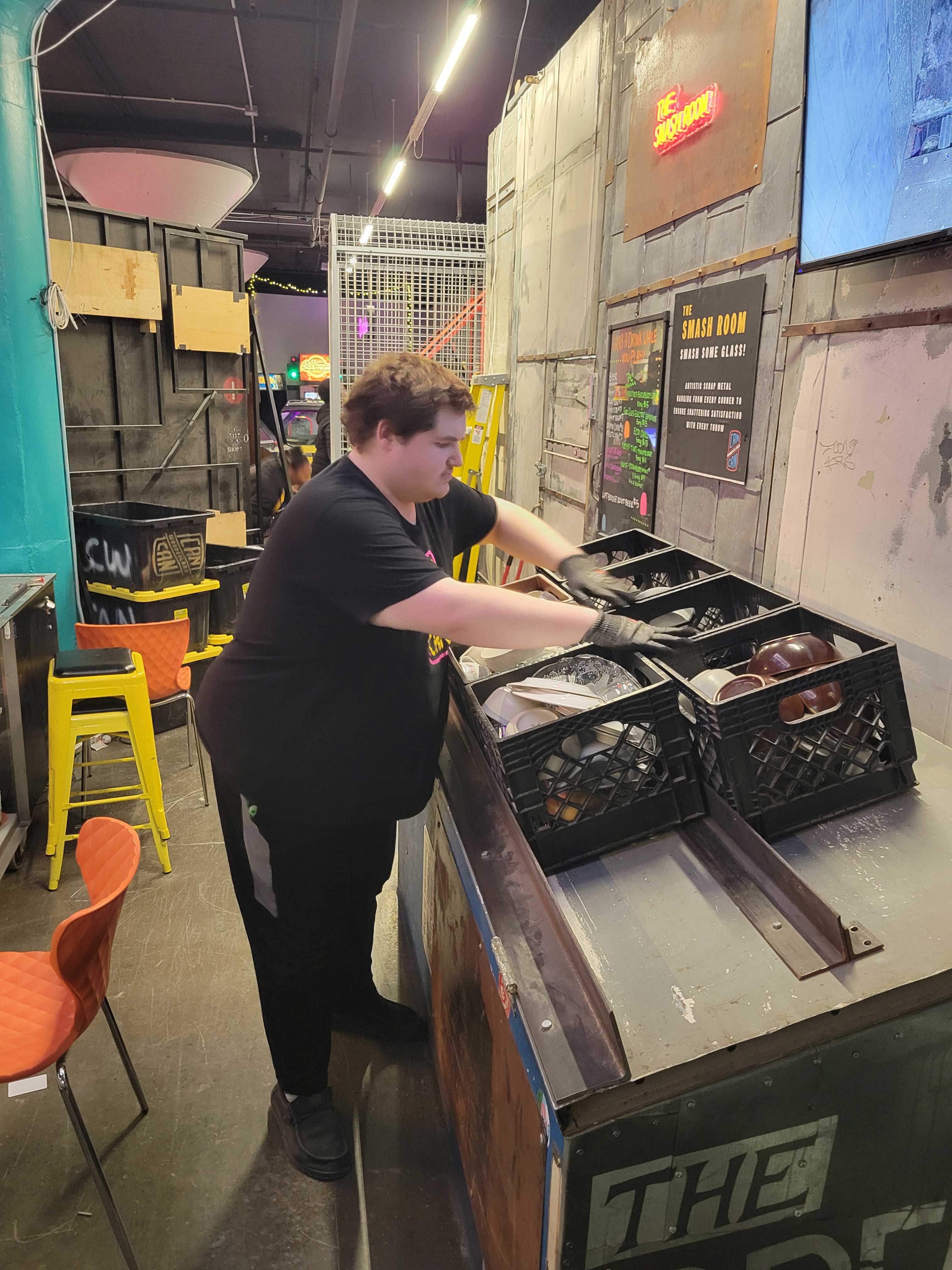Young man wearing black shirt and pants stands in an industrial room loading items into brown milk crates lined up on metal shelves