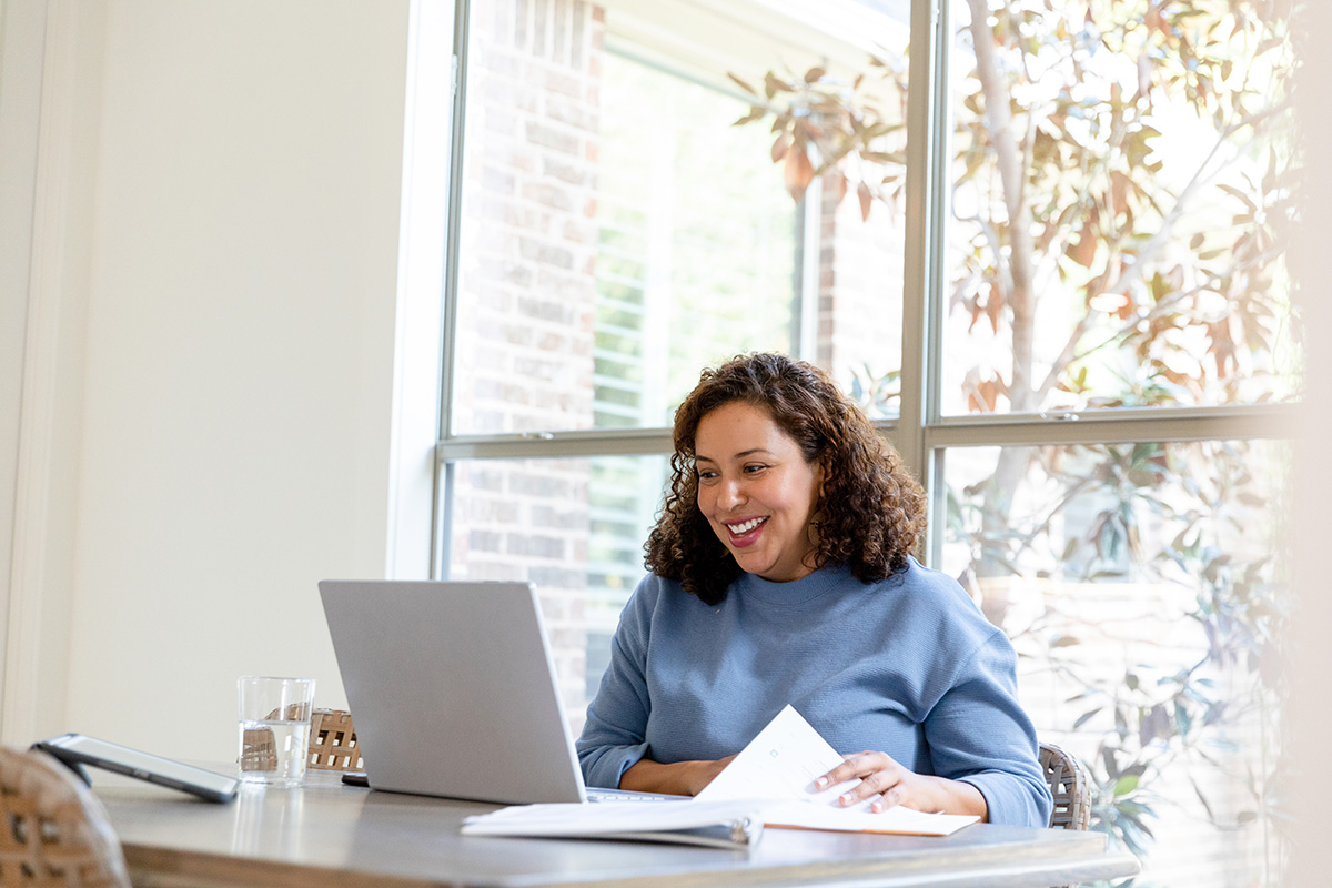 Woman with curly hair and light blue sweater sits at a table in front of a large window. A laptop is open in front of her