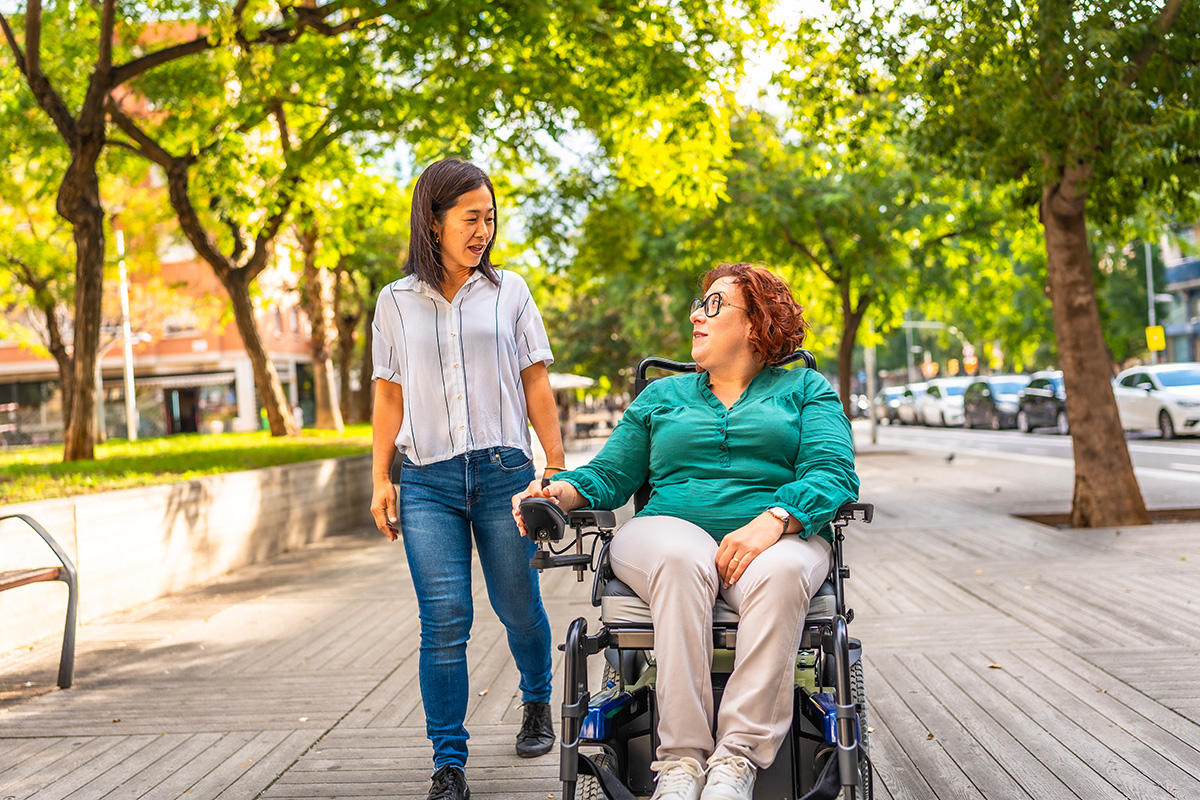 A middle aged woman with red hair, wearing a green blouse and khakis, uses a motorize wheelchair on a city sidewalk. A young Asian woman walks beside her, wearing a white blouse and jeans