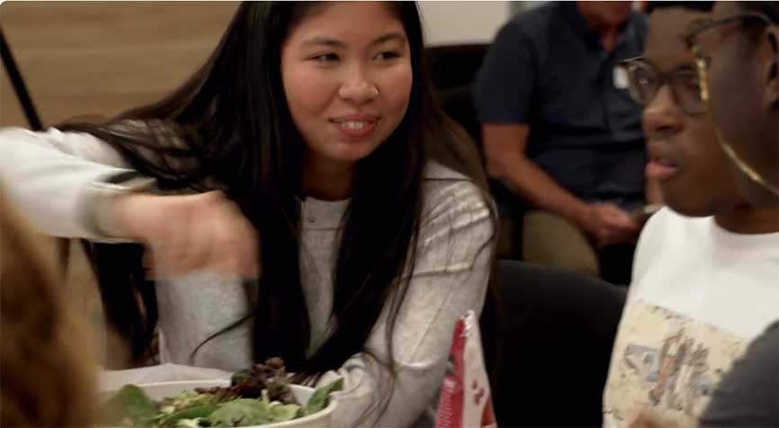 Asian woman in white sweater tosses a large salad while two seated African American men look on