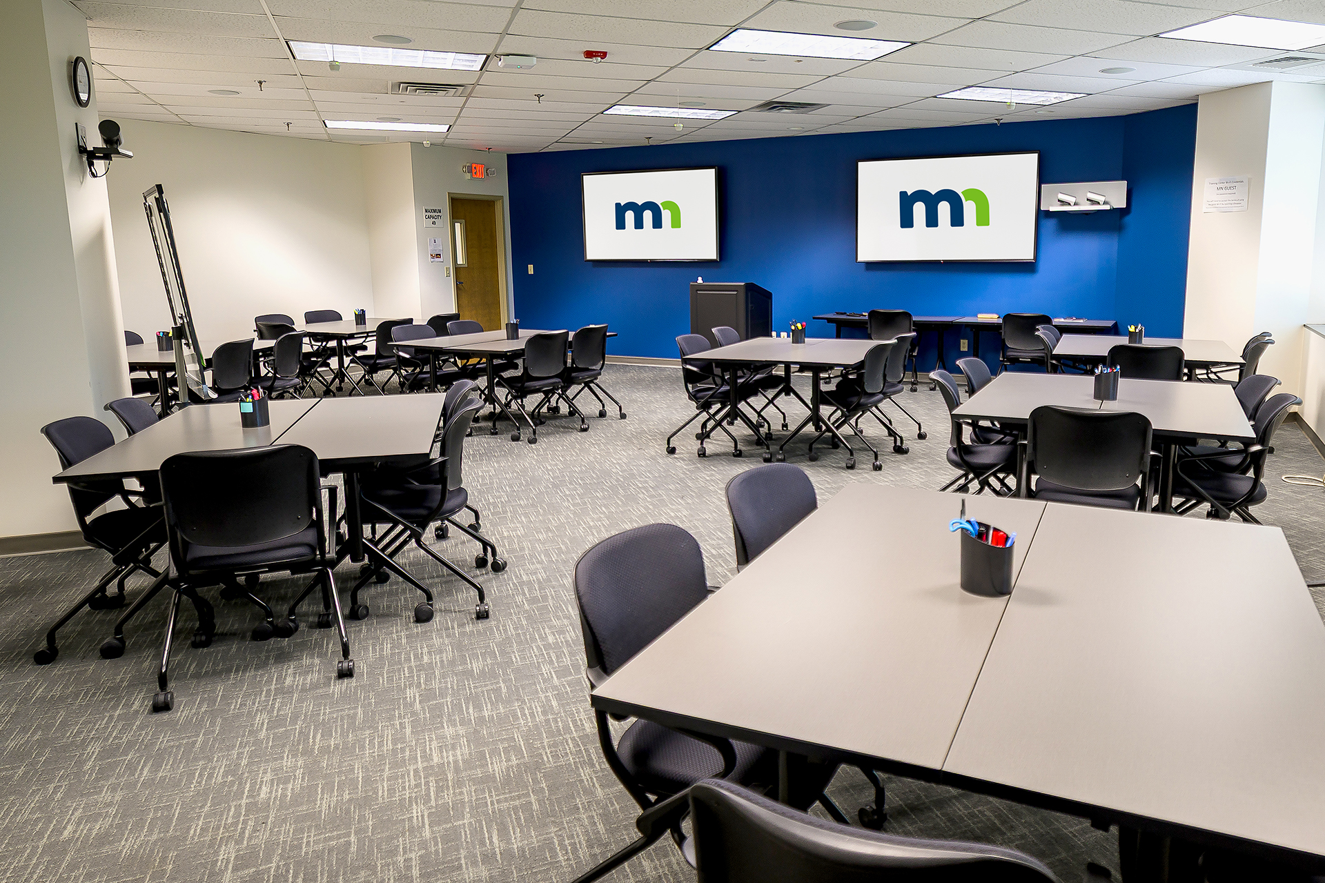 Aristotle room at the Training Center with a front lectern, several tables, and two TVs for presentations