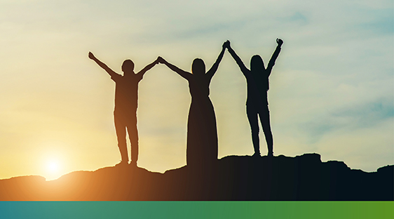 Three people standing on a rocky hill, holding hands with their arms and hands in the air, and the sunrise in the background.