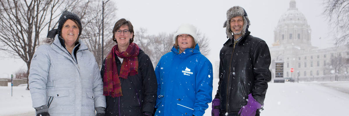 Snow falling as Kim, Kris, JoAnn, and Jay stand in front of the Minnesota State Capitol Building.