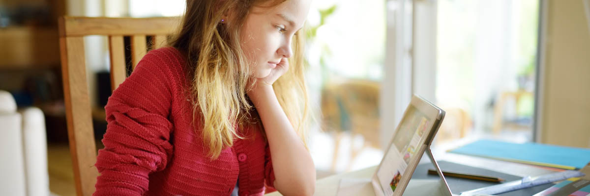 A girl reading a digital tablet and writing in a notebook.