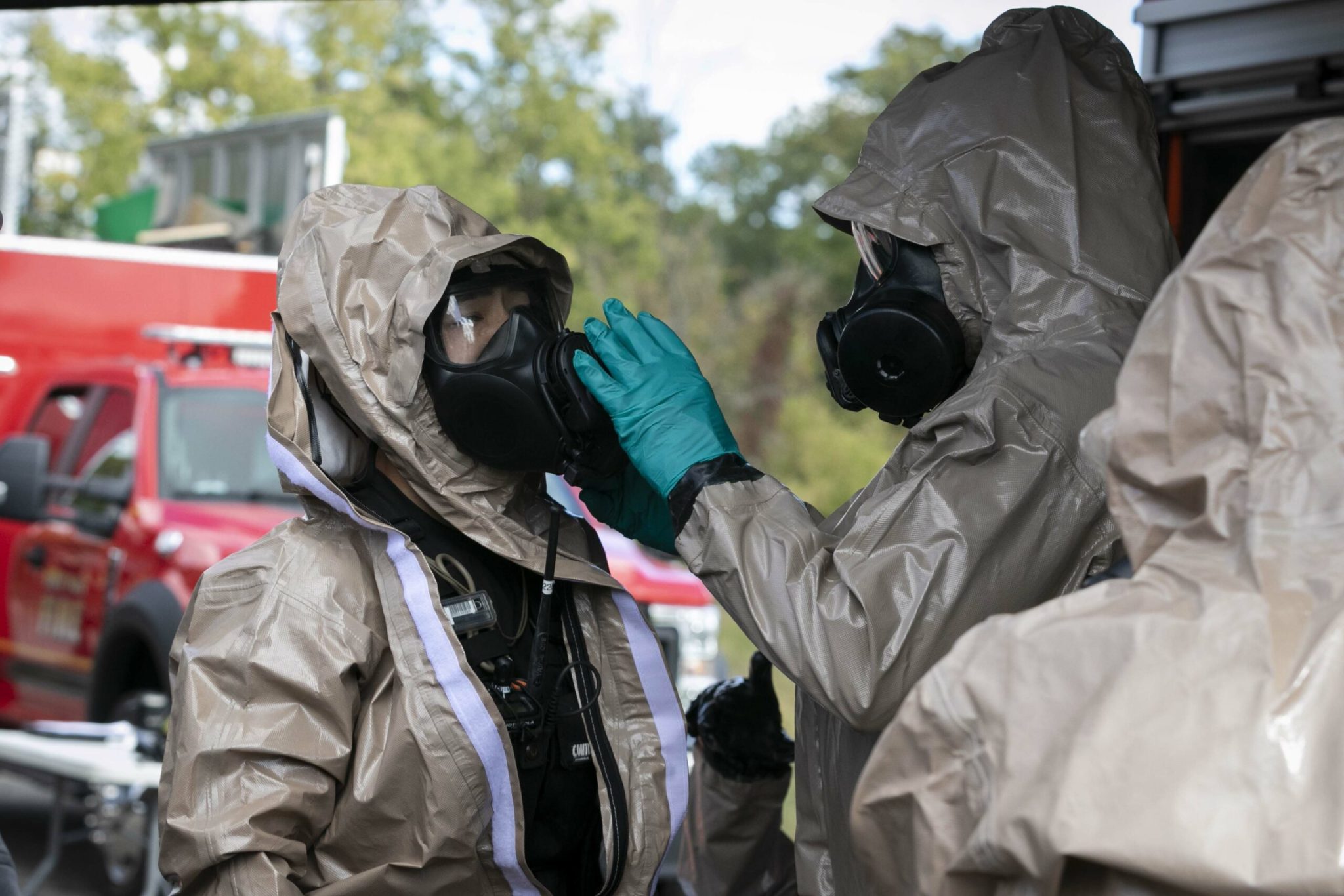 Two people in hazmat suits, and one is helping another get their gas mask situated